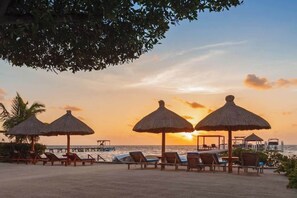 A beach with thatched umbrellas and lounge chairs, a pier, and boats in the background during sunset.