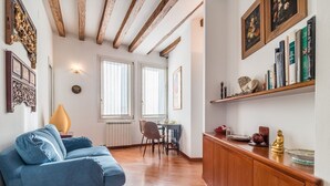 A cozy living room with a blue sofa, wooden beams on the ceiling, a dining area with a table and chairs, and a bookshelf with books and decorative items. - 7 WINDOWS ON VENICE APARTMENT (Venezia)