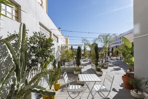 Un espace extérieur aménagé en terrasse, avec des meubles en métal blanc, des plantes en pot et un ciel d’un bleu limpide.
