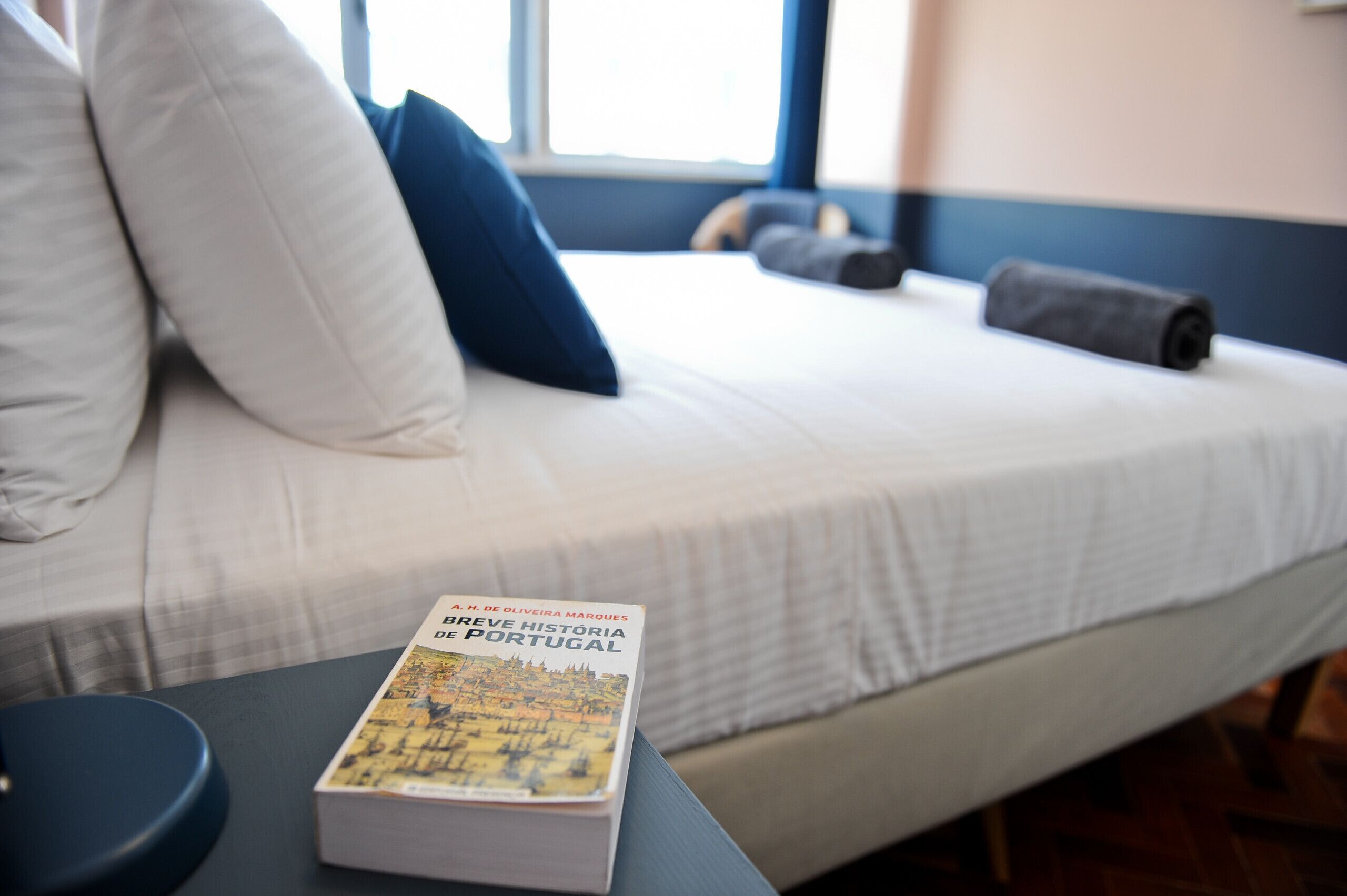 Una cama cuidadosamente hecha con un libro titulado "Breve História de Portugal" sobre una mesita de noche.