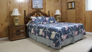 A bedroom with a wooden paneled wall, a bed with a floral bedspread, two bedside tables with lamps, and a framed picture above the bed. - Private, Beautiful & Quiet Guesthouse On The Withlacoochee River (Yankeetown)
