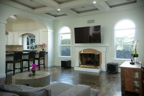 A spacious living room with a fireplace, large windows, and a kitchen area in the background.