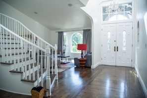A spacious foyer with a wooden staircase, a chandelier, and a large window with curtains.