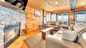 A cozy living room with a stone fireplace, wooden ceiling, and large windows offering a view. - Rivers Ledge Cabin with breathtaking views, hot tub, & indoor pool! (Bryant)