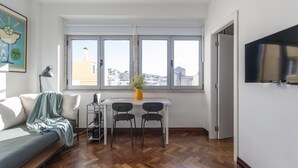 Una sala de estar moderna con un sofá, una mesa de comedor con sillas, una planta en maceta y un ventanal con vistas a la ciudad.