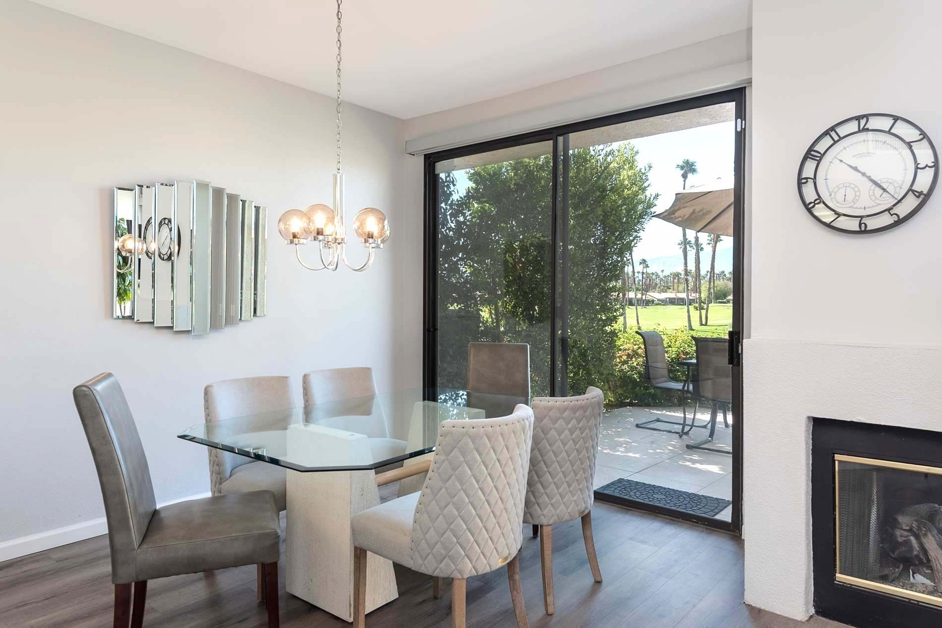 A dining area with a glass table and cushioned chairs, a chandelier, a wall clock, and a view of a golf course through a sliding glass door.