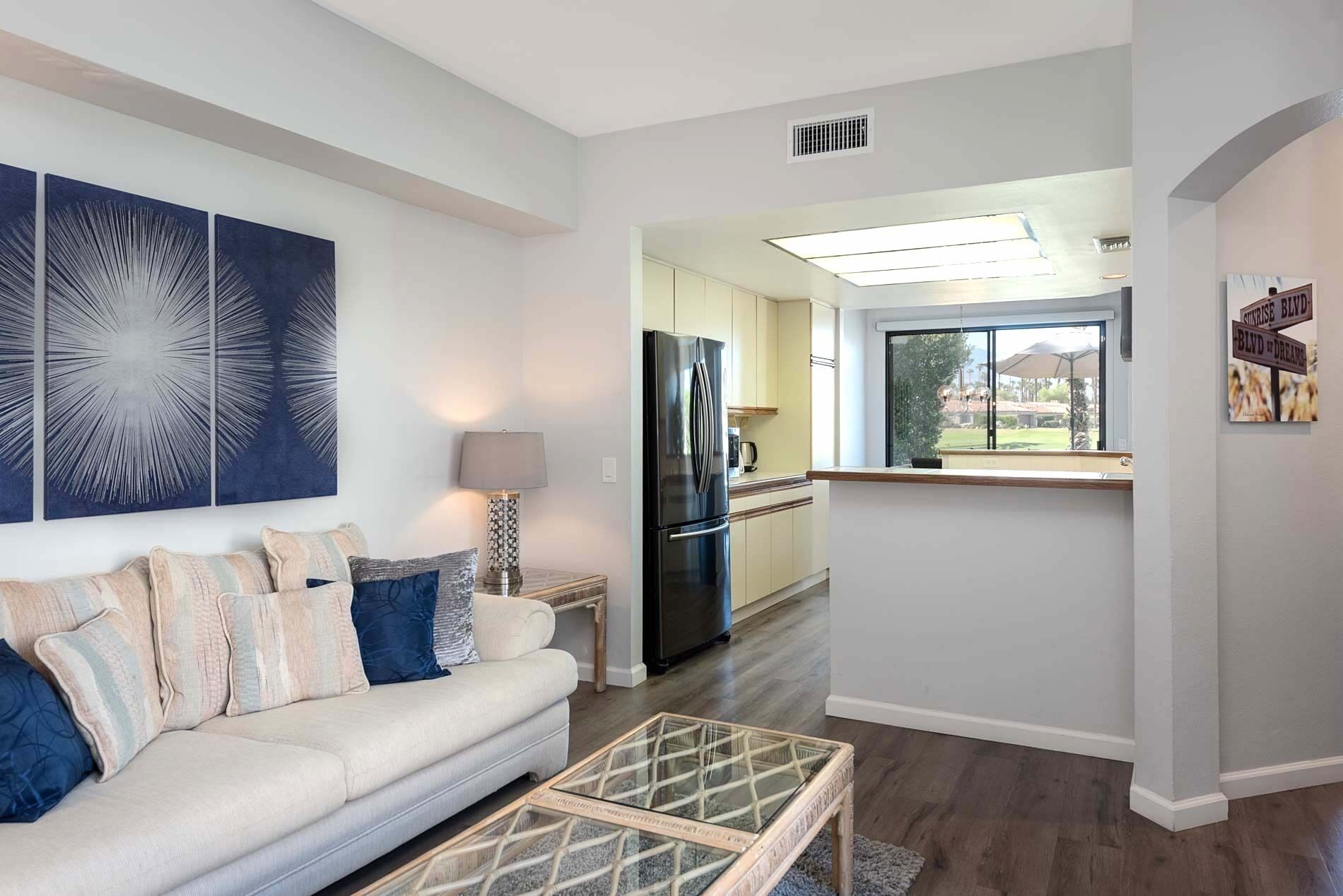 A modern living room with a sofa, a coffee table, and a kitchen area with a refrigerator and a window.