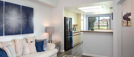 A modern living room with a sofa, a coffee table, and a kitchen area with a refrigerator and a window.