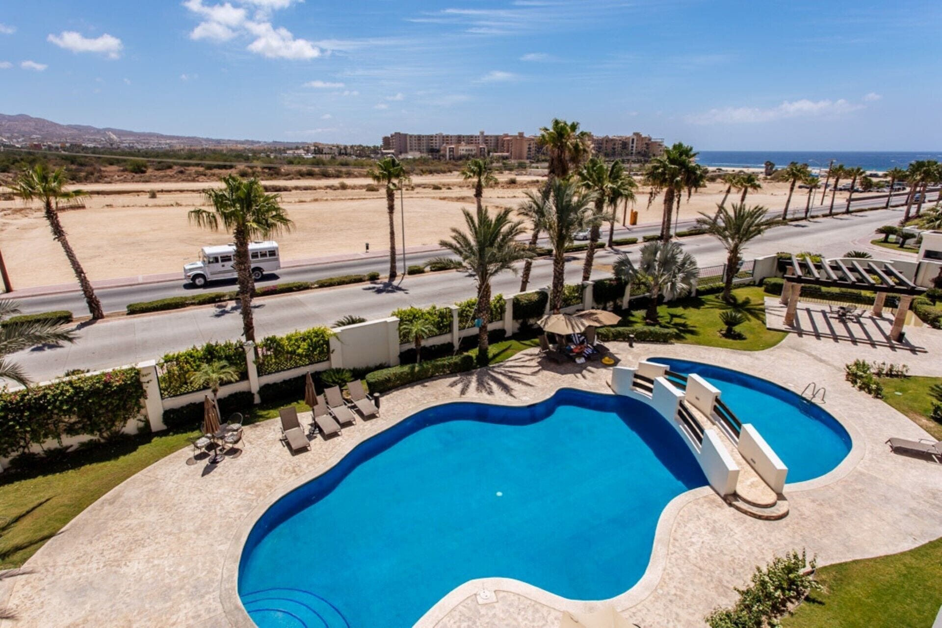 A swimming pool with lounge chairs, palm trees, and a view of a beach and buildings in the background.