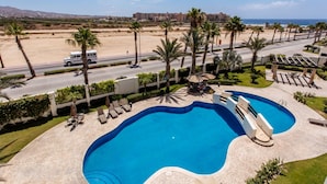 Ein Swimmingpool mit Liegestühlen, Palmen und Blick auf Strand und Gebäude im Hintergrund.