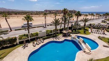 A swimming pool with lounge chairs, palm trees, and a view of a beach and buildings in the background.