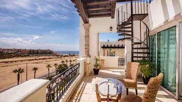 A balcony with wicker chairs, a glass table, and a view of the beach and buildings.