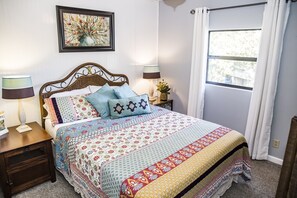 Une chambre à coucher avec un lit, deux tables de chevet avec des lampes, une fenêtre avec des rideaux et une œuvre d’art encadrée accrochée au mur.