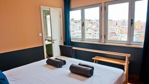 A hotel room with a bed, a wooden bench, a window with a city view, and a door.
