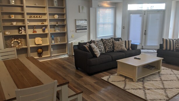 A modern living room with a dark-colored sofa, a white coffee table, and a wooden dining table. There is a large white shelving unit filled with various decorative items. - DOWNTOWN NEWPORT HOTSPOT! Private Parking included - Open floor plan, spacious! (Newport)