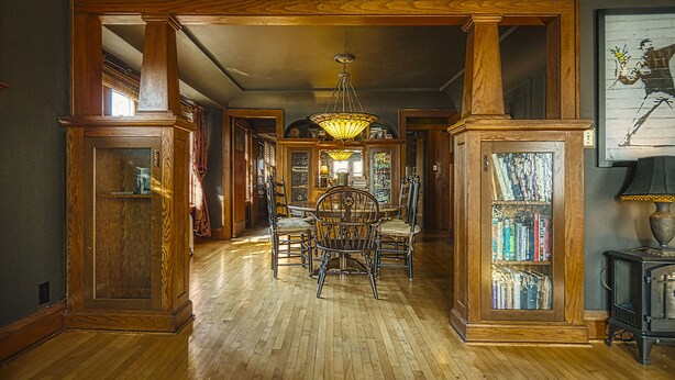 A spacious hallway with wooden flooring, a central dining table with chairs, a chandelier, and a bookshelf with books.