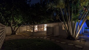 Ein modernes Haus mit hellem Innenraum, umgeben von einem Garten mit Palmen und einem Weg zum Eingang.