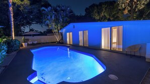 Ein Pool mit ungewöhnlicher Form, umgeben von einem Haus mit großen Fenstern und einer Terrasse mit Liege und Sonnenschirm.
