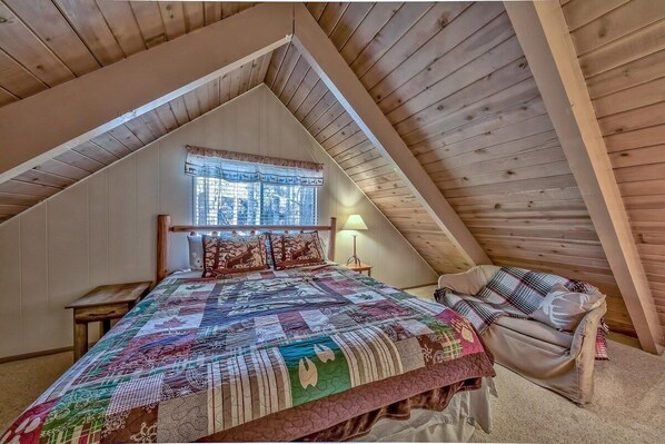 Ein Schlafzimmer mit schrägen Wänden, einem Bett mit Steppdecke, einem Fenster mit Vorhängen, einem Nachttisch und einem Sessel.