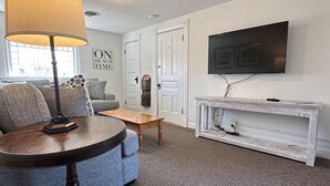 A living room with a round table, a sofa, a coffee table, a TV on a white stand, and a door with a sign.