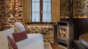 Une pièce chaleureuse avec un mur en pierre, une fenêtre en bois avec des rideaux, un lit avec une literie blanche et des oreillers rouges, et un poêle à bois.