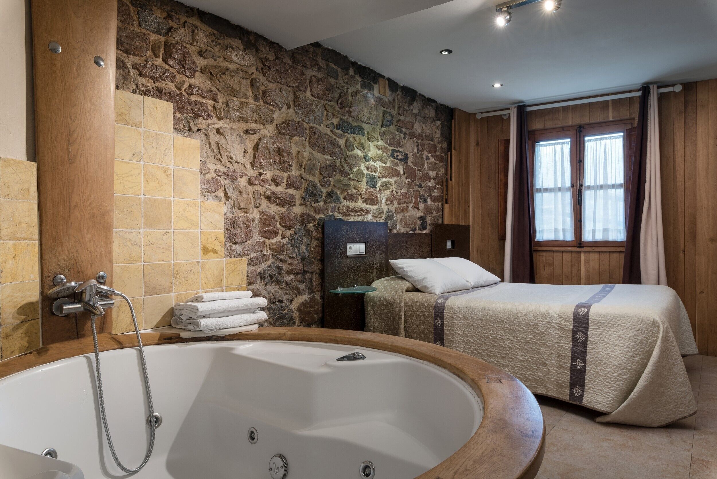 Ein Zimmer mit Steinwand, einer großen Badewanne, einem Bett mit gesteppter Bettdecke und einem Nachttisch mit Lampe.