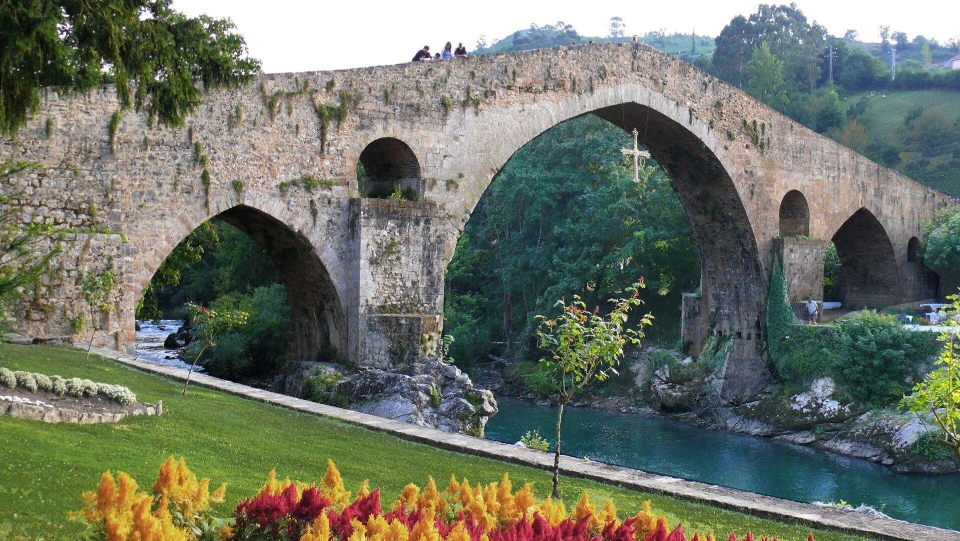 A stone bridge with multiple arches over a river, surrounded by greenery and a well-maintained lawn.