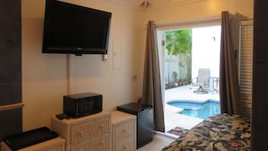 Una habitación con televisión, un refrigerador pequeño, un aparador de mimbre, una cama con cubrecama floral y vistas a una piscina y mobiliario de patio.