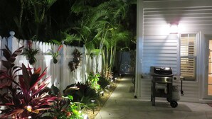 Un sendero bordeado de plantas diversas que conduce a una casa blanca con una parrilla de barbacoa en el exterior.