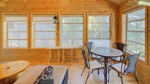 Interior de una cabaña de madera con una mesa de comedor y sillas, un sofá y ventanas amplias.