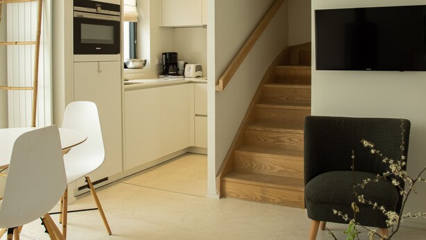 A modern kitchen with white cabinets, a wooden staircase, a dining area with a white table and chairs, and a black armchair with a vase of flowers. - MV 2 - L'Éveil de la Côte - House by the beach (Audresselles)