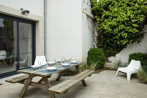 Eine Terrasse im Freien mit einem Holztisch, gedeckt für ein Essen, umgeben von weißen Plastikstühlen und einem Betonboden.