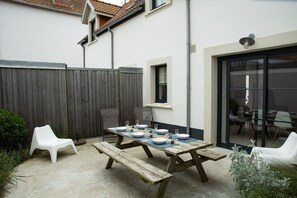 Eine Terrasse im Freien mit einem Holztisch, der für das Essen gedeckt ist, umgeben von weißen Stühlen und einer Holzbank.