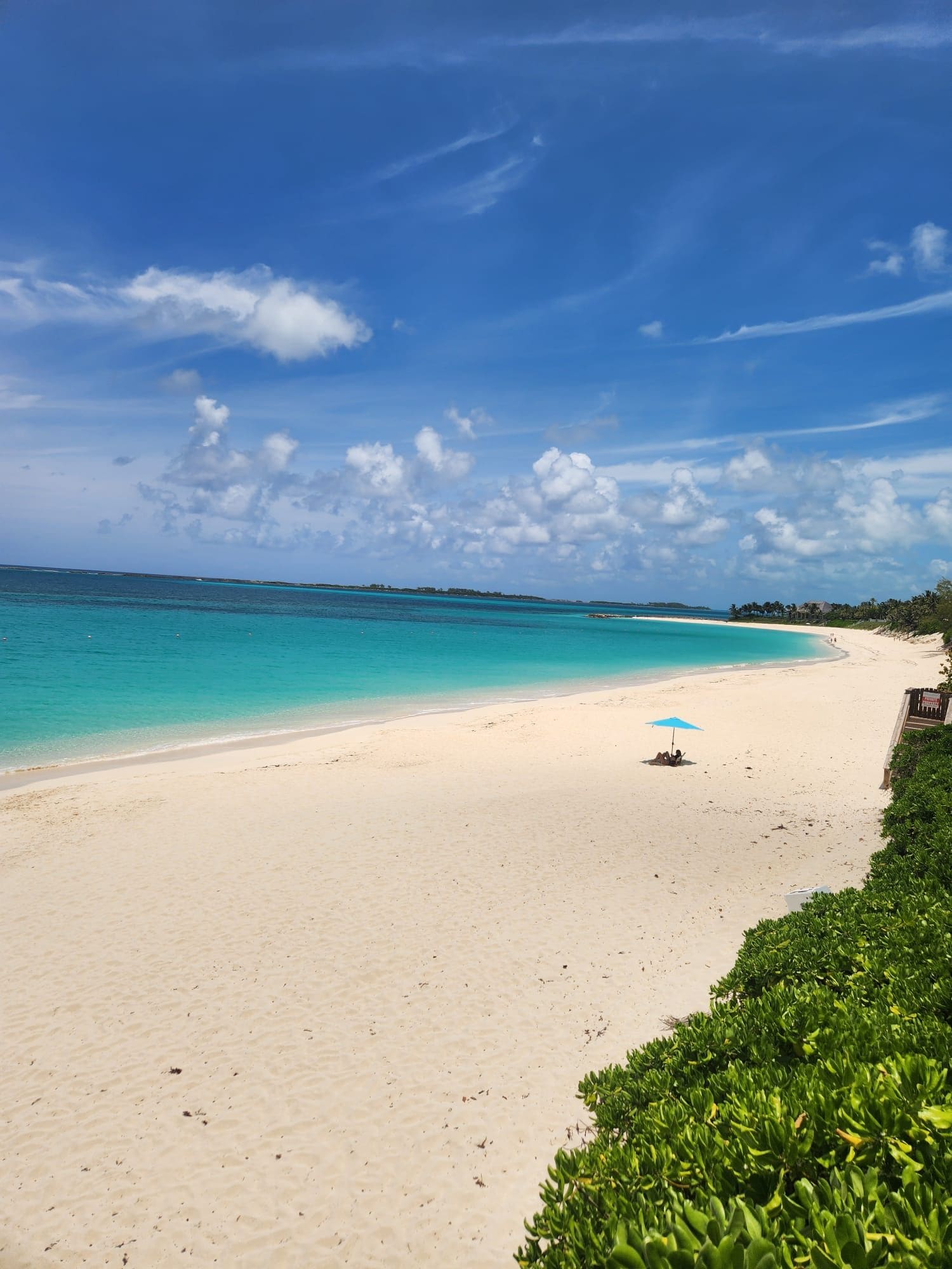 A beach with clear turquoise water, a sandy shore, and a few people under a blue umbrella.