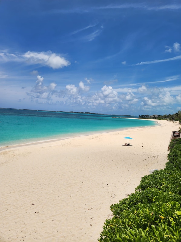 A beach with clear turquoise water, a sandy shore, and a few people under a blue umbrella.