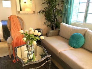 Un soggiorno con un divano, un tavolo con un vaso di fiori, una poltrona con una coperta, una pianta grande e una finestra con tende.