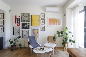 A modern living room with a wooden floor, two white armchairs, a coffee table with books, and various framed artworks on the walls.
