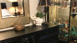 A room with a large mirror, a black dresser with decorative items, a lamp, and a traditional tapestry.