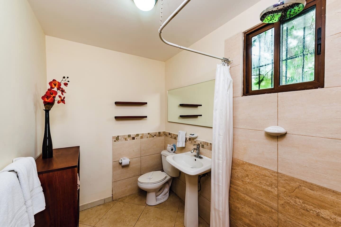 A bathroom with a toilet, sink, and shower. There is a vase with flowers on a wooden table, and a window with a view of greenery.