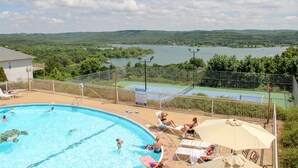 Una piscina con gente, cancha de tenis y vistas a un lago y colinas.