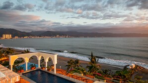 Ein Hotelpool mit Blick auf den Ozean und den Strand, umgeben von Palmen und einer Bergkulisse.