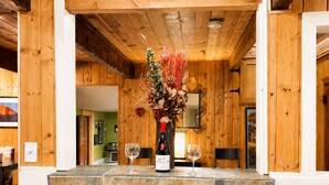 Interior de madera con barra, dos sillas, una botella de vino y un florero con plantas secas.