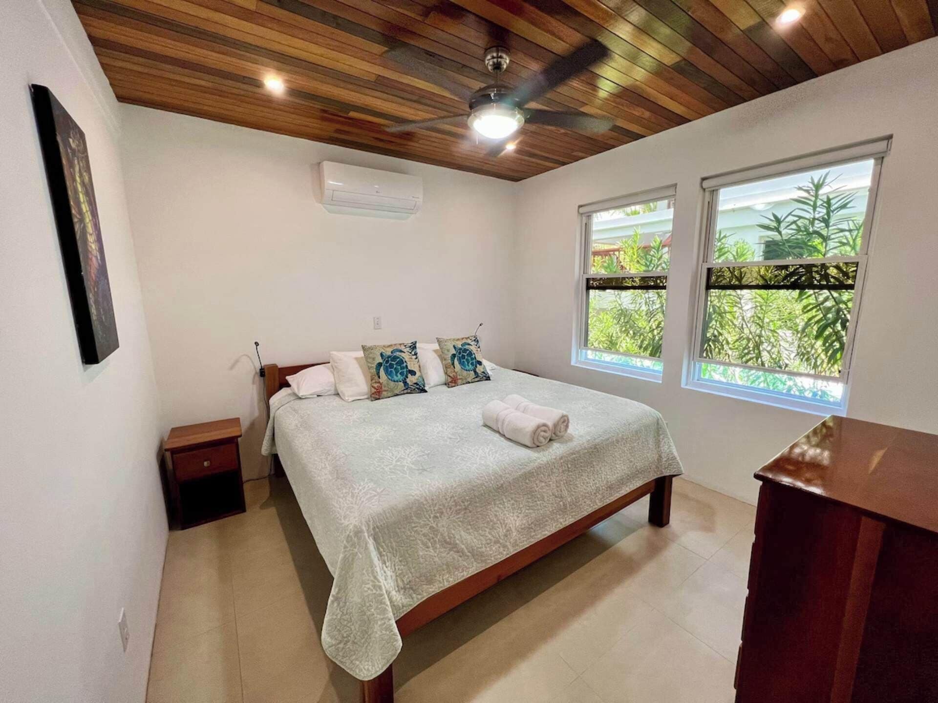 A bedroom with a wooden ceiling, a bed with a white bedspread, two windows with a view of greenery, and an air conditioning unit on the wall.