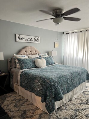 A bedroom with a tufted headboard, a bed with patterned bedding, a ceiling fan, a wall with motivational decor, and a window with sheer curtains.