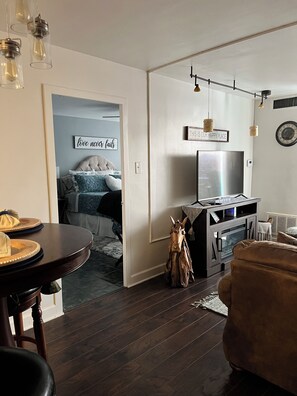 Un salon moderne avec un parquet en bois foncé, un canapé en cuir marron, une table à manger avec des chaises, un téléviseur et une cheminée. Un panneau est accroché au mur avec l’inscription suivante : « ICI, NOUS SOMMES HEUREUX ».