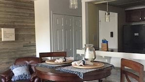 Un comedor con una mesa redonda de madera, dos sillas y lámparas colgantes. Se ve una cocina al fondo.