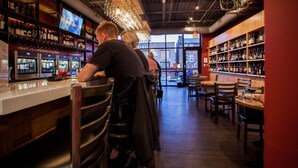 A bar area with shelves of bottles, a TV screen, and a view of the street outside.