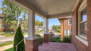 A covered porch with brick pillars and a metal railing, overlooking a street with parked cars and residential buildings.