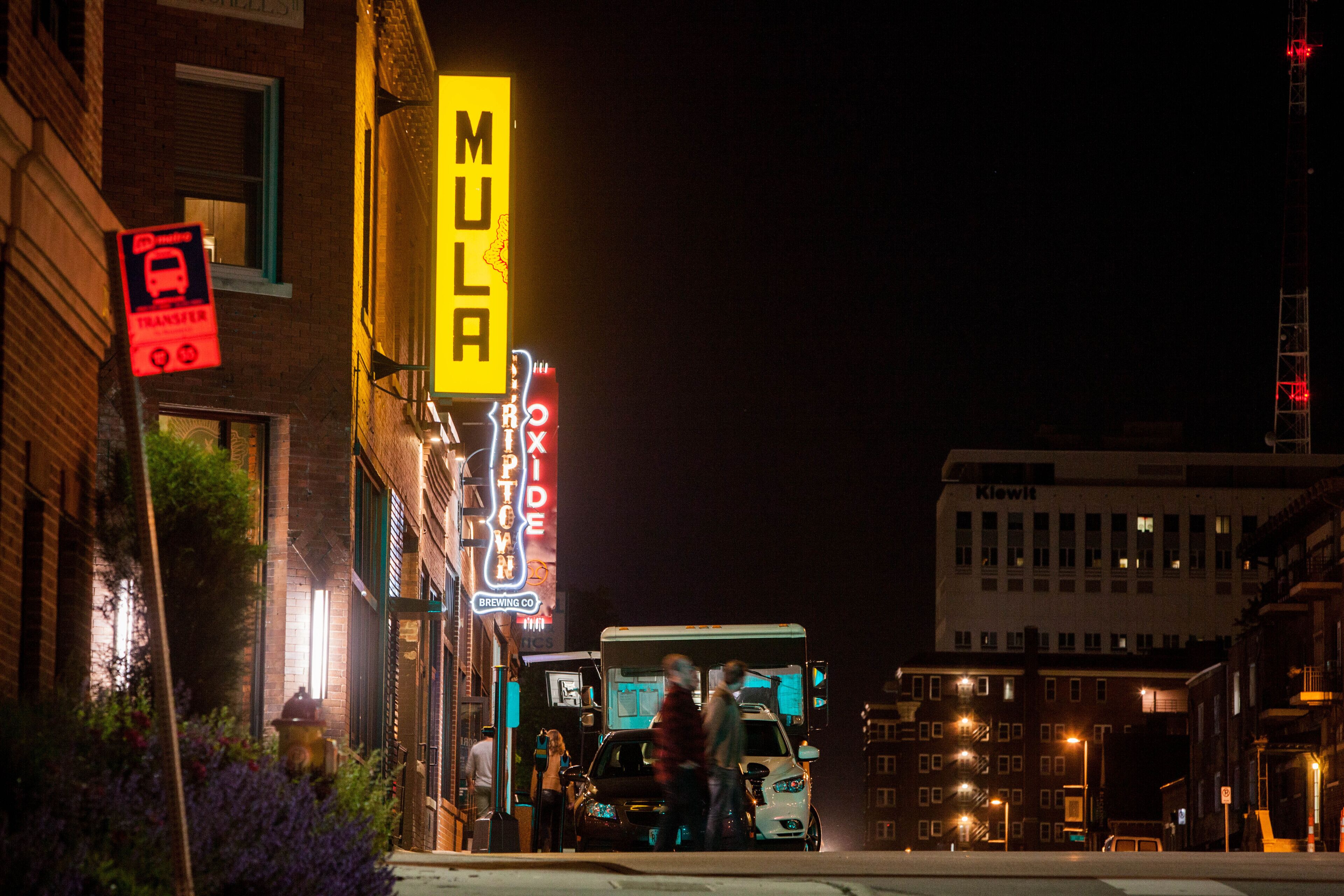 A nighttime urban street scene with a prominent yellow neon sign for 'MULA' and a 'Brewing Co' sign.