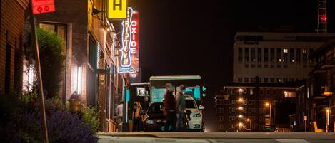 A nighttime urban street scene with a prominent yellow neon sign for 'MULA' and a 'Brewing Co' sign.
